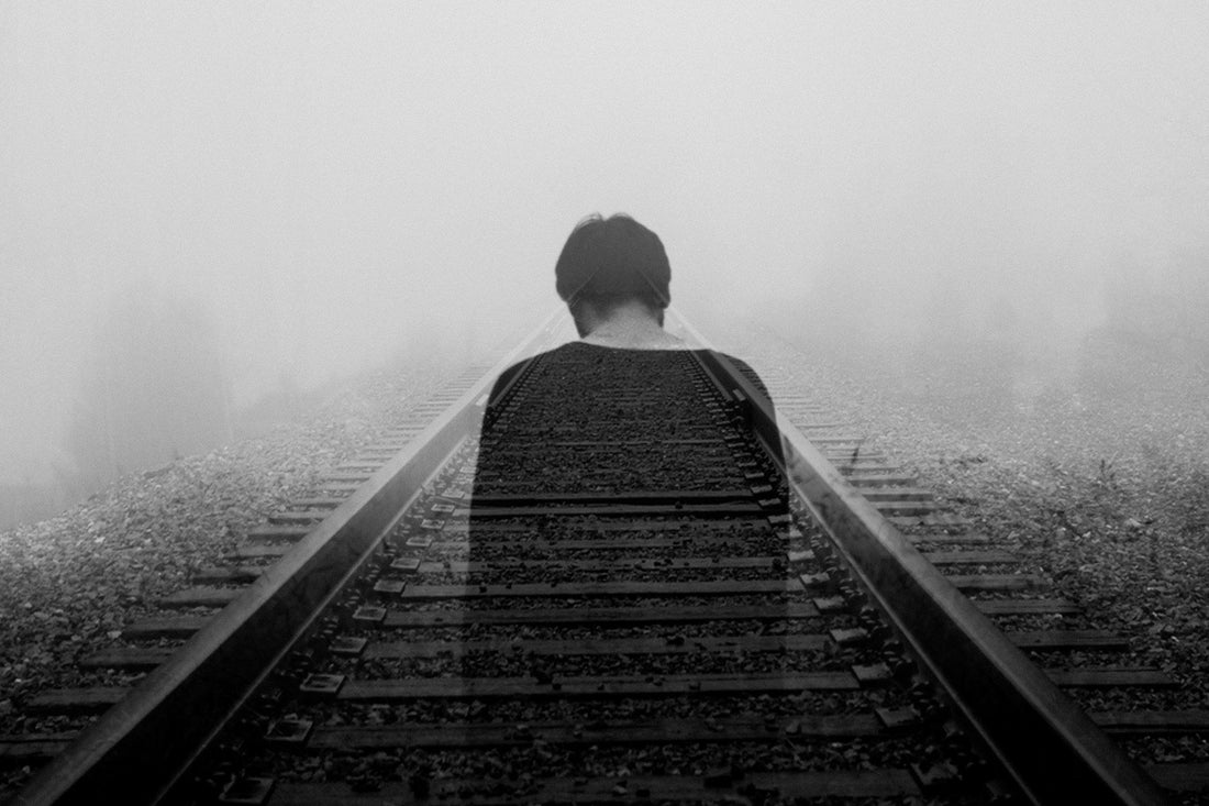 Black and white photograph of a solitary person sitting on empty train tracks disappearing into thick fog, representing the uncertainty and isolation of choosing to be alone versus staying in a fading or unfulfilling relationship