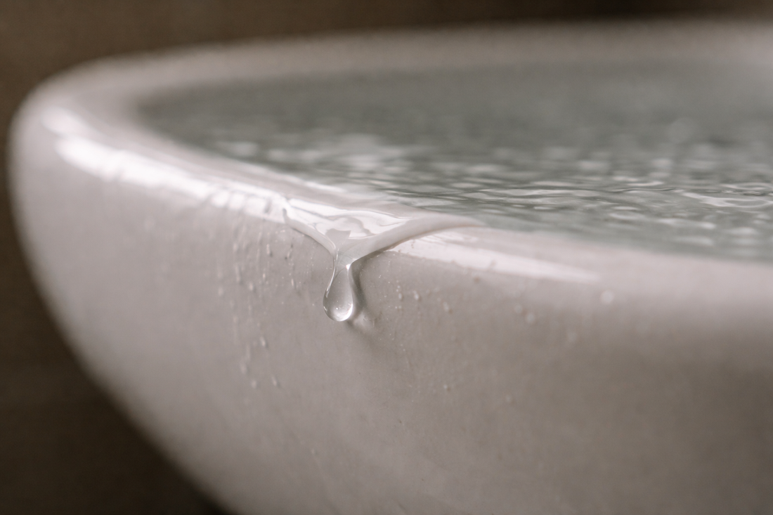 Extreme macro side profile of a white ceramic sink filled to the brim, water forming a slight convex meniscus with a single droplet hanging at the edge in soft natural light.