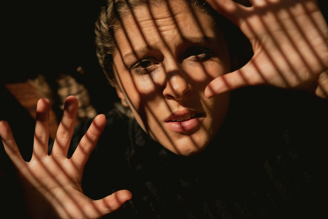 Woman shielding her face behind raised hands and window-blind shadows, symbolizing fear, confusion, and emotional control in a narcissistic marriage.