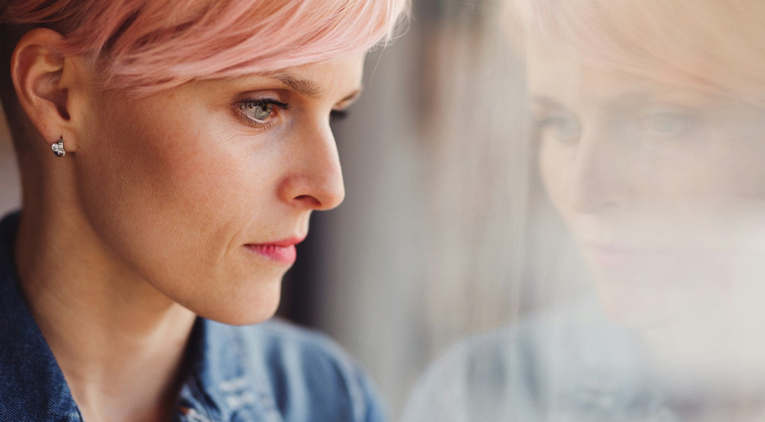 Woman with pink hair looking thoughtfully through a window, reflecting breakup comparison and wondering if an ex is happier with someone new.