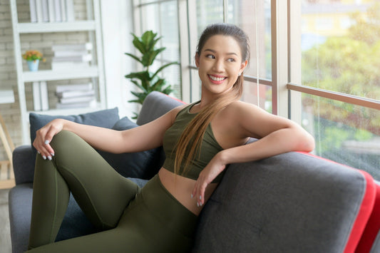 Woman wearing dark green fitted activewear while seated on a sofa, illustrating shapewear and front-smoothing clothing designed to reduce visible camel toe