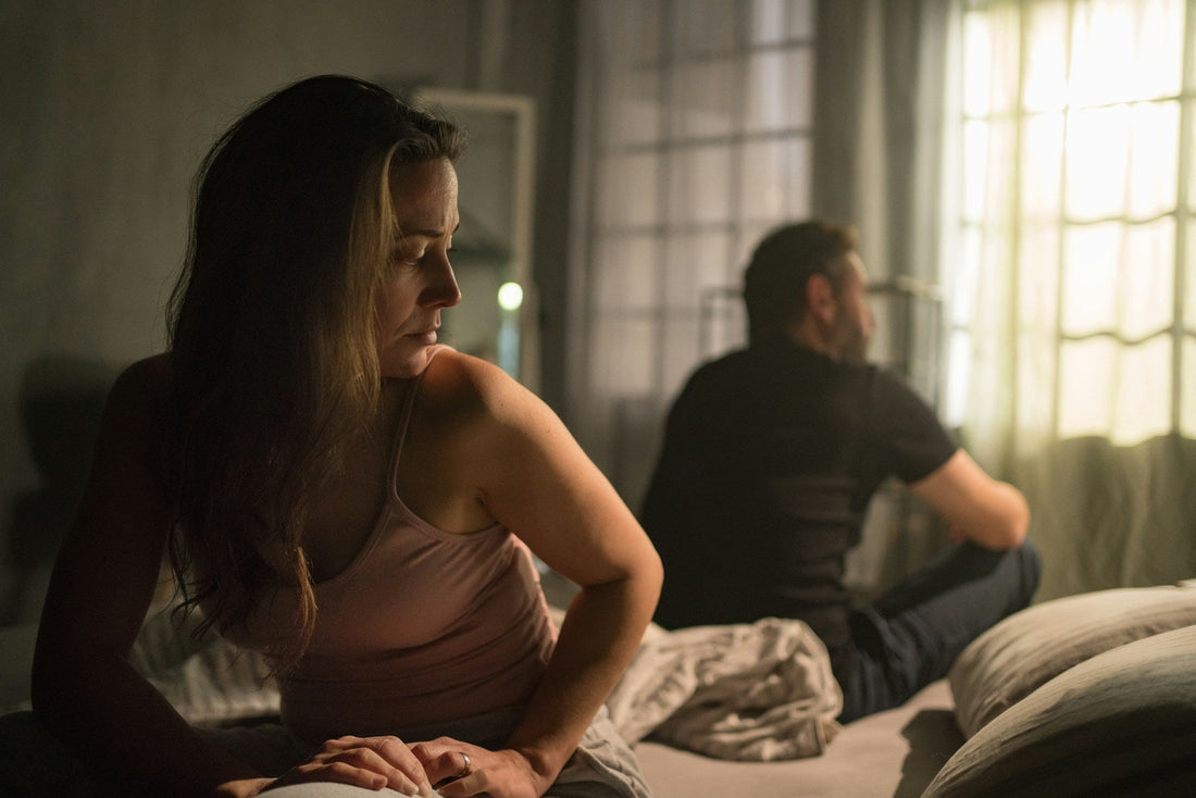 A woman sits on a bed in a dimly lit bedroom, looking off-camera with a sad expression, while a man sits on the bed in the background facing a bright window.