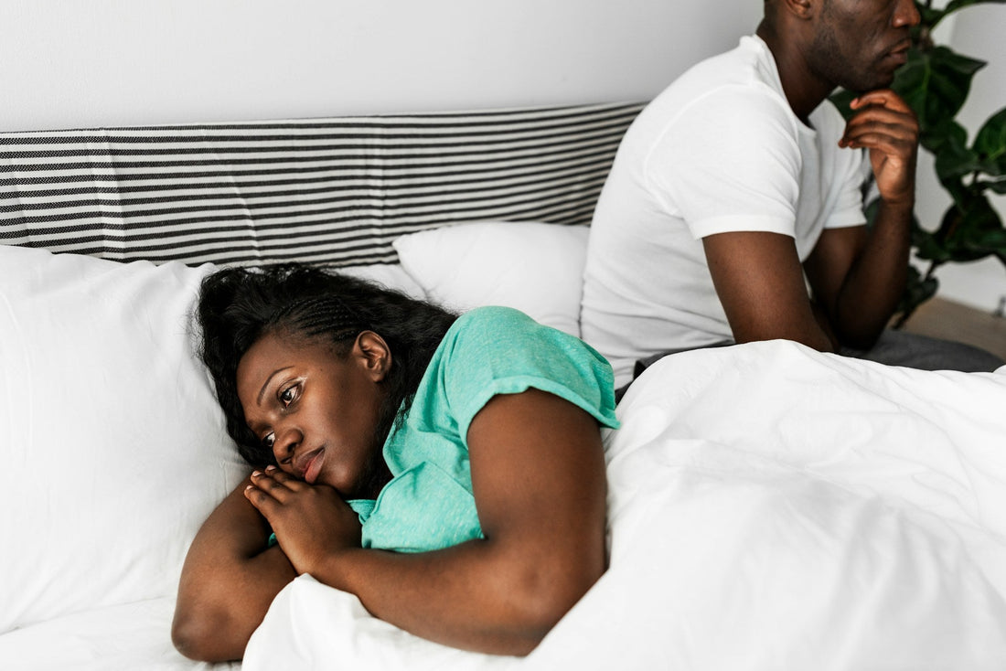 A couple in a bedroom, with a woman lying in bed looking up at a man who is sitting on the edge of the bed, wearing a white t-shirt and a mint green shirt.