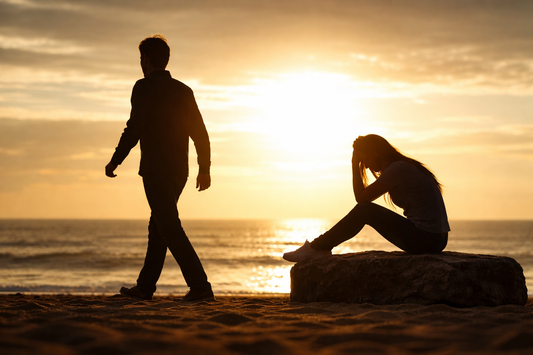Silhouetted couple at sunset on a beach, one person walking away while the other sits alone in emotional distress after a breakup