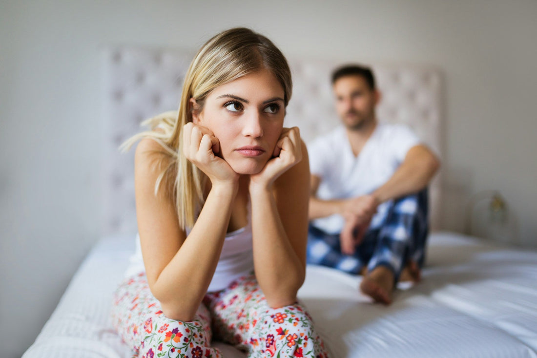 A blonde woman in a bedroom sits on a bed with her head resting on her hands, looking off-camera with a sad, thoughtful expression. A man in a white shirt and blue plaid pajama pants sits blurred in the background on the bed.