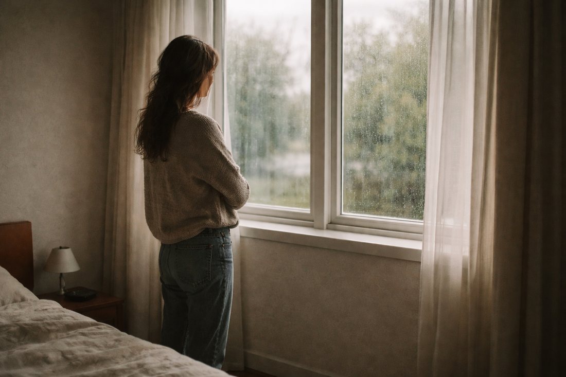 person standing quietly by a rain-covered window in soft natural light reflecting on the uneven emotional process of healing after a breakup