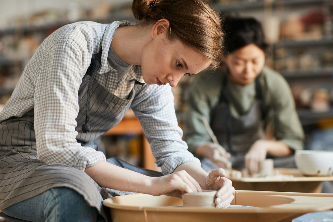 young woman sitting at pottery wheel and adjusting edges of clay vessel while making pot in workshop enjoying her new hobby