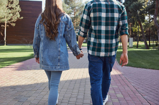 A couple walking hand-in-hand away from the camera on a paved path in a park. The woman wears a light blue denim jacket and jeans, and the man wears a green and white plaid button-up shirt with blue jeans.