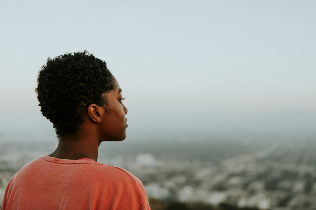 A woman with short curly black hair is shown from the side in profile, wearing a coral-pink t-shirt and looking out over a hazy cityscape at dusk.