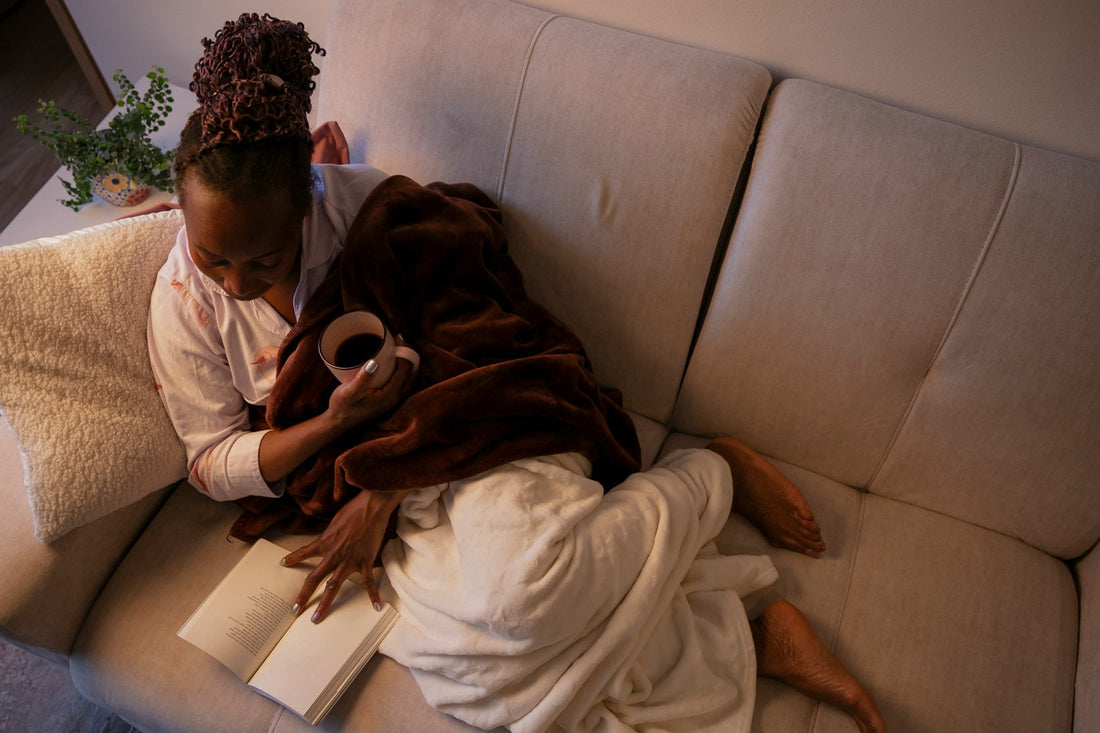 Woman relaxing alone on a sofa with a book and warm drink, representing healthy alone time, personal space and independence within a relationship.