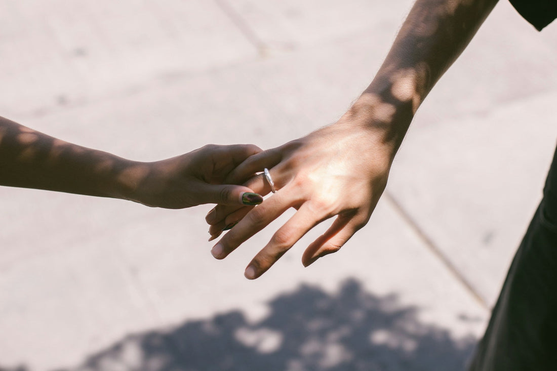 A close-up outdoor shot of two hands reaching toward each other, with a thin silver ring on one finger. The background is a sunlit concrete surface with dappled shadows.