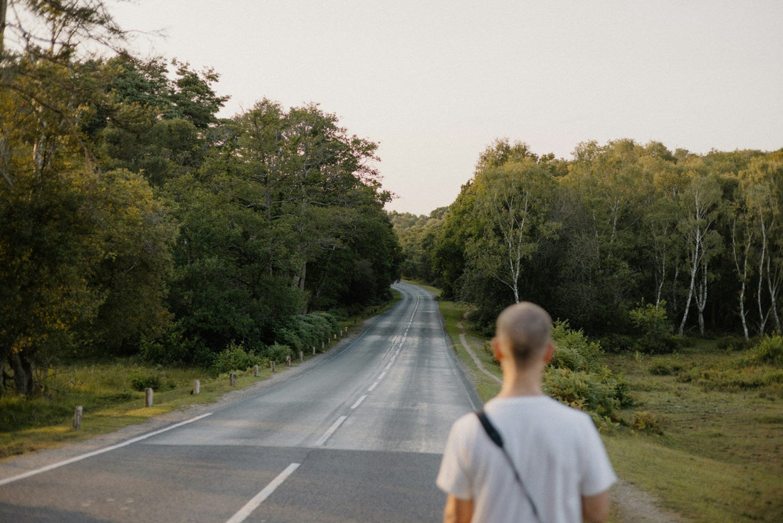 A man in a white t-shirt stands on the side of a paved road surrounded by dense green trees, viewed from behind as he looks down the road.