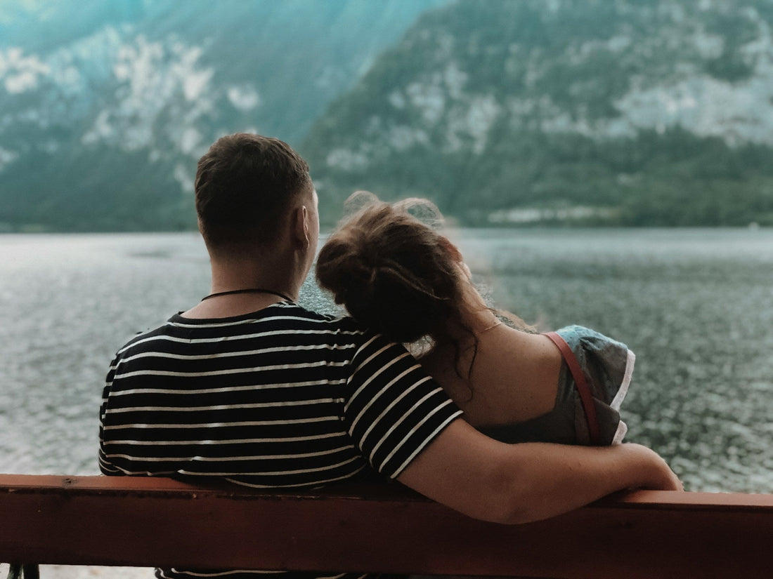 A man and a woman sit on a wooden railing, viewed from behind, looking out over a body of water with mountains in the background. The man wears a black and white striped t-shirt, and the woman rests her head on his shoulder.