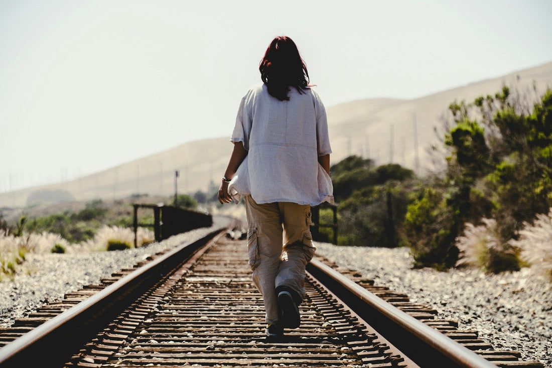 A person seen from behind walking down railroad tracks in an outdoor landscape with hills, shrubs, and gravel under a bright sky.