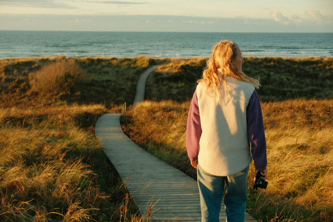 A woman with blonde hair stands on a wooden boardwalk path in a grassy dune field, looking out toward the ocean at sunset. 