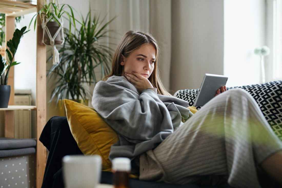 A woman relaxes on a patterned couch in a cozy living room, wrapped in a gray blanket and holding a tablet. She is wearing beige knit pajama pants and has a yellow pillow behind her.