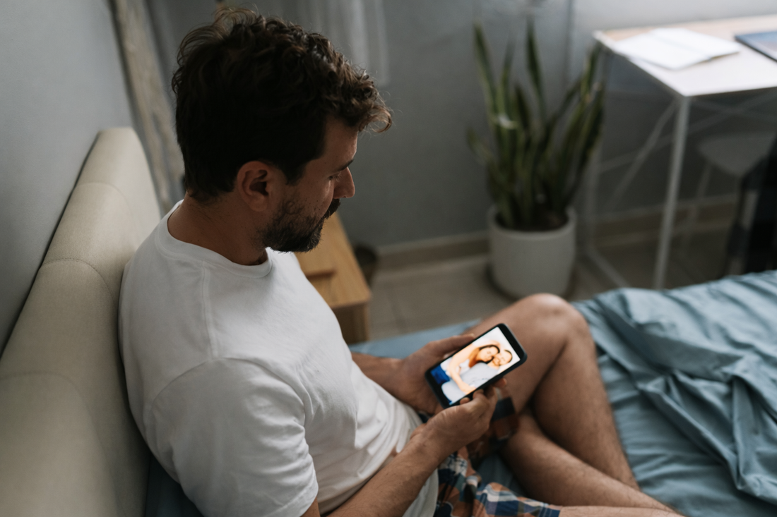 Man sitting on a bed in a softly lit room, looking down at his phone in a quiet, reflective moment.
