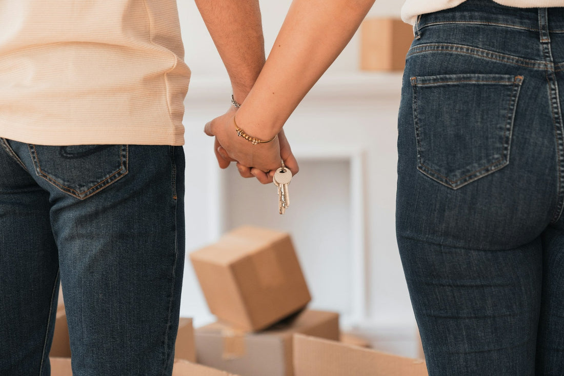 Two people standing arm in arm, viewed from behind, wearing blue jeans and holding a set of keys, moving in after long distance relationship. Cardboard boxes are visible on the floor in a bright indoor setting.