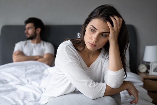 Woman sitting on bed looking worried while her partner sits in the background, representing relationship doubt, anxiety, and constant questioning in a romantic relationship.