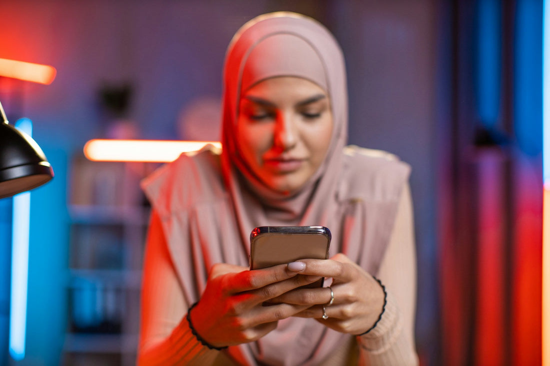 Muslim woman in a hijab looking at her phone in a private setting, representing the emotional pull of a secret relationship that is difficult to leave.