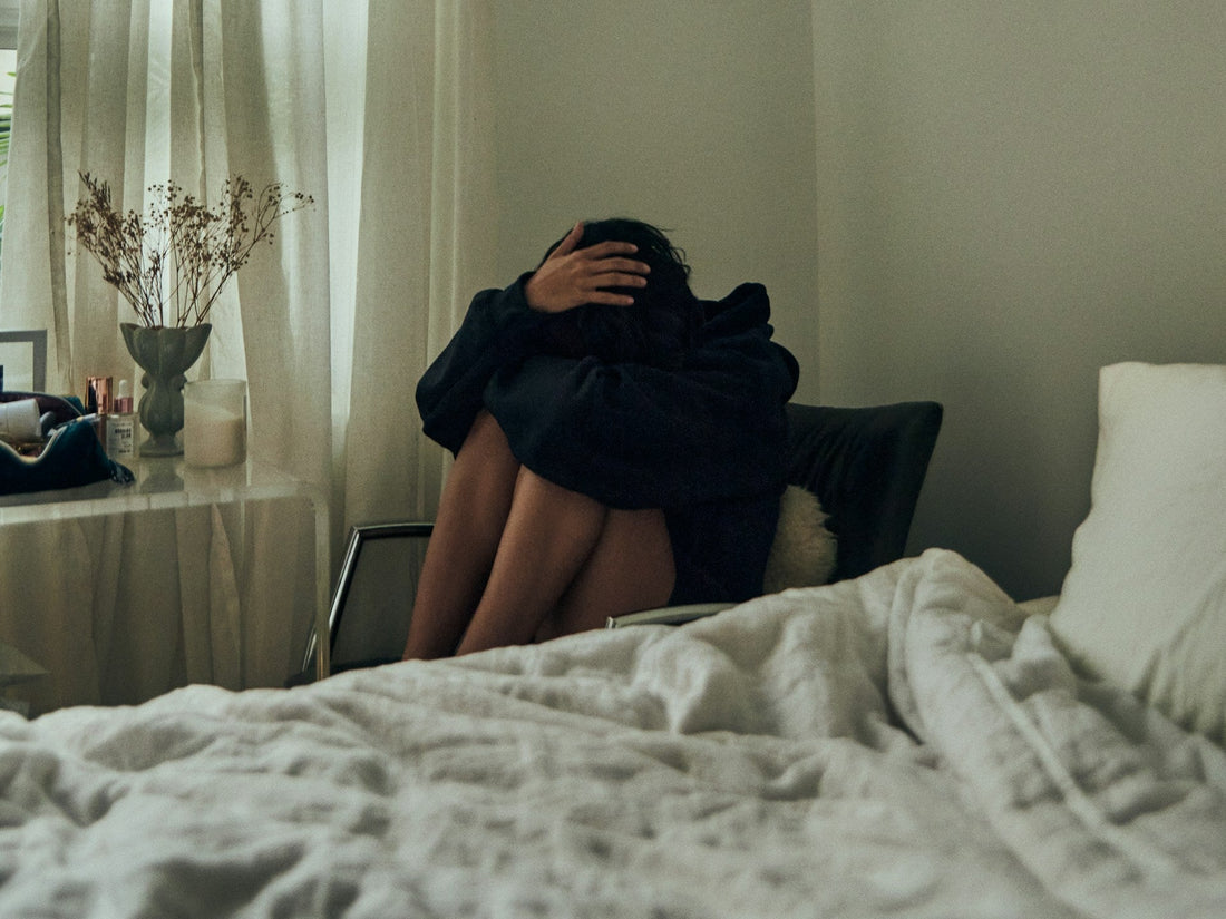 A person in a black outfit sits curled up on a chair in a dimly lit bedroom, with a bed and white curtains in the foreground.