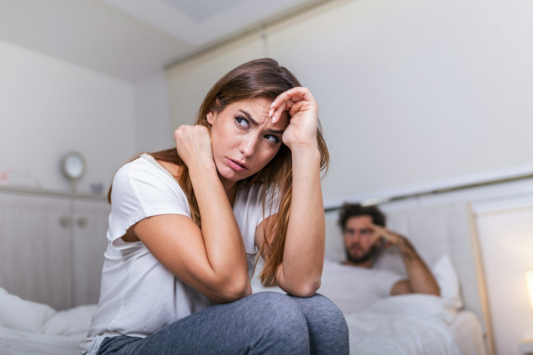 A woman sits on a bed in a bedroom, looking distressed with her hands on her face, while a man sits blurred in the background on the bed.
