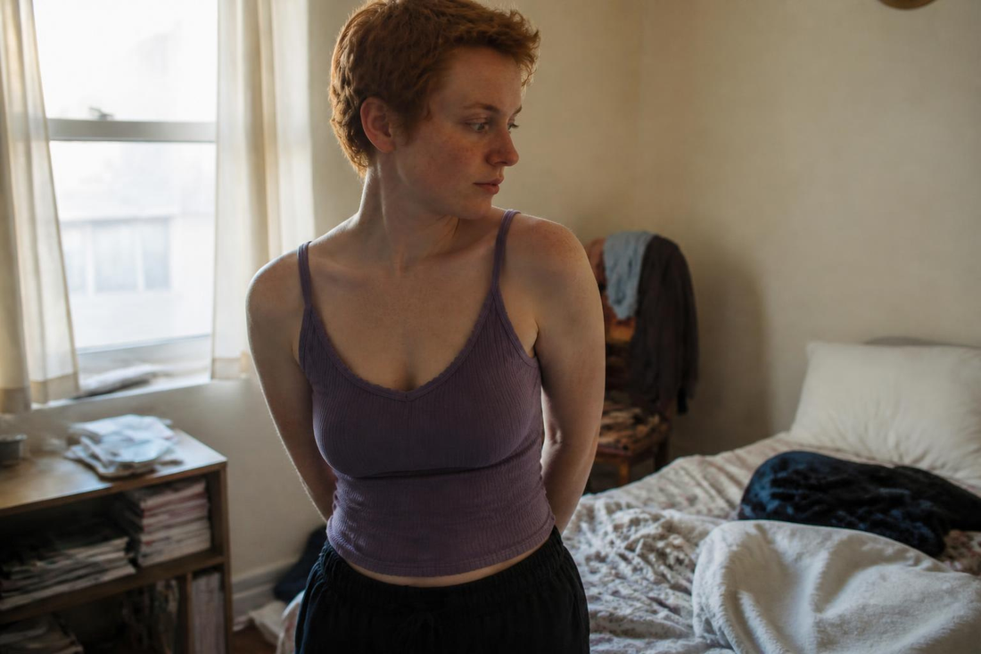 Woman with short red hair and freckles looking thoughtfully at her body in a softly lit, lived-in bedroom, illustrating body insecurity and sexual confidence.