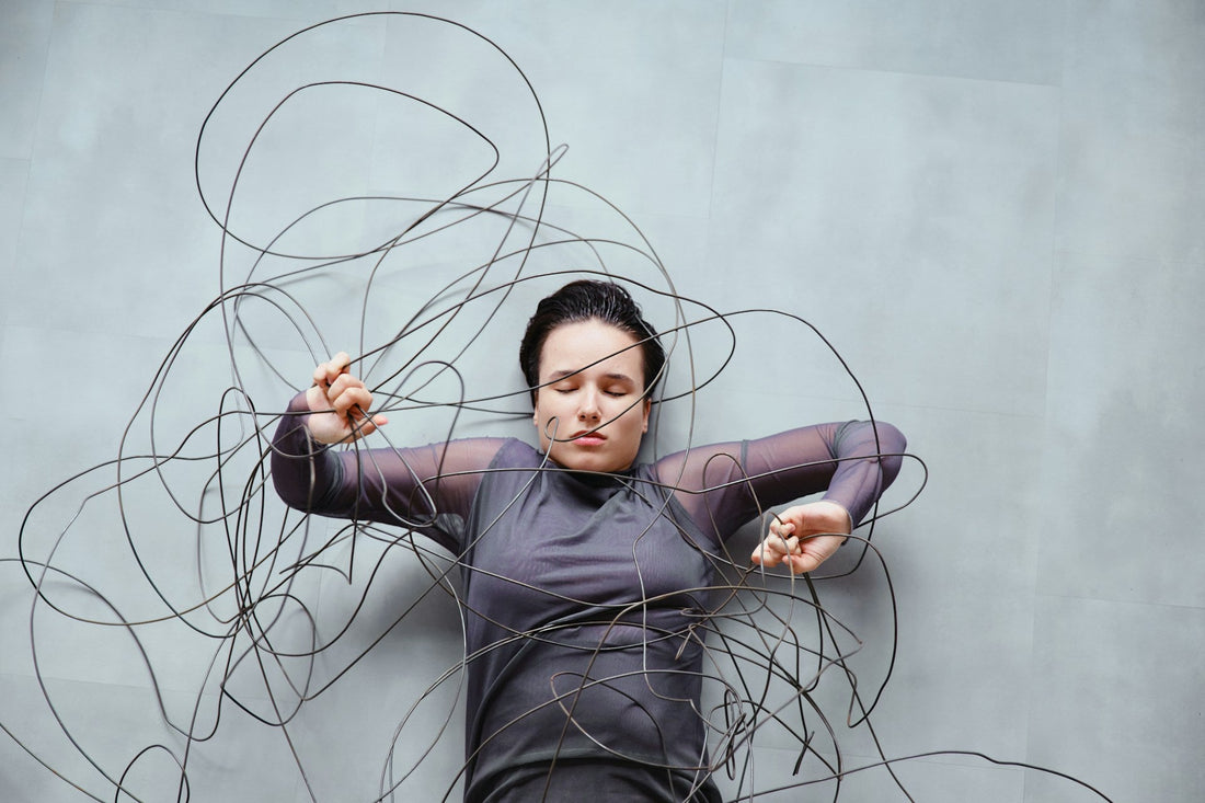 A woman lying on a gray concrete floor surrounded by tangled black cables, wearing a dark gray long-sleeve top.