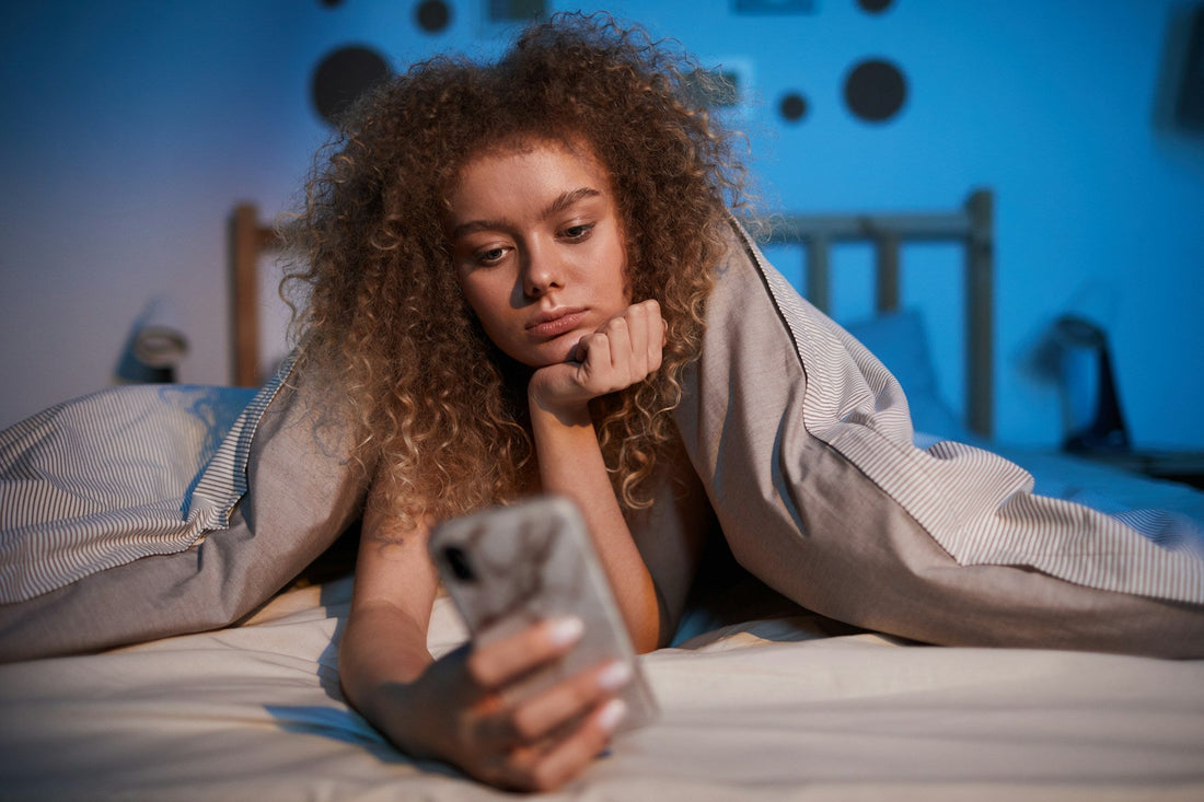 Woman lying in bed looking uncertain at her phone while deciding whether to send a second text after getting no reply.