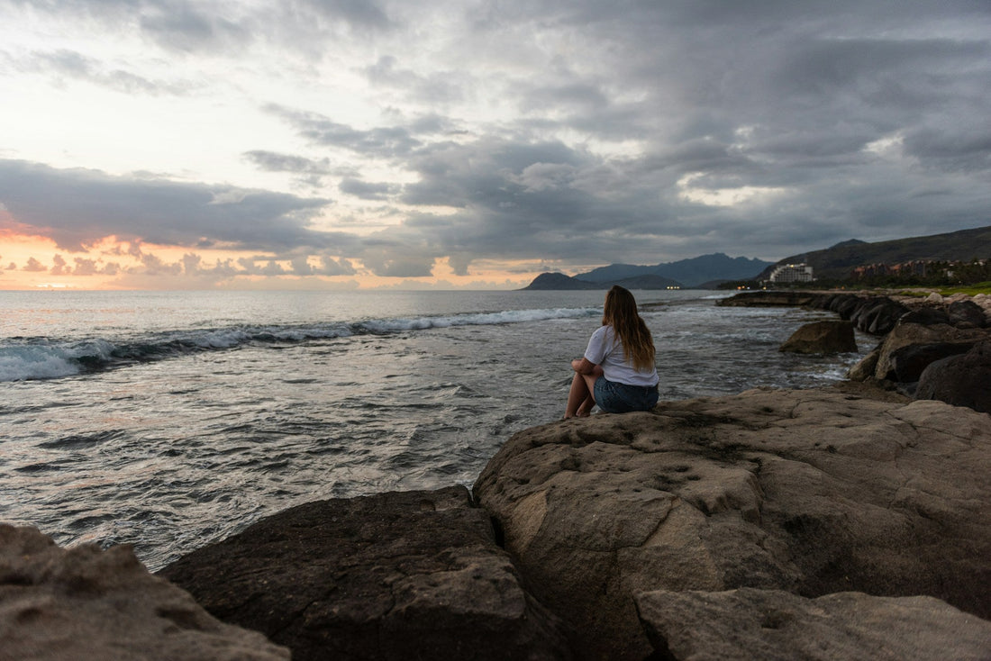 Woman sitting alone on rocks beside the ocean at sunset, reflecting calm, self-trust, and healing from self-abandonment.