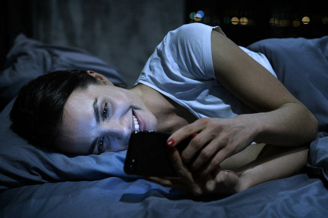 Woman smiling while lying in bed and looking at her phone, illustrating playful long-distance relationship chatting and romantic connection.
