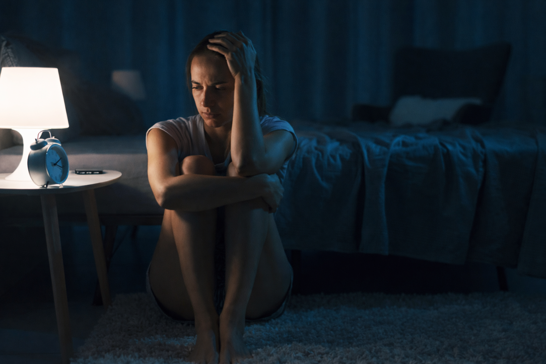 Woman sitting on the floor beside her bed at night, arms wrapped around her knees, looking down in quiet sadness while a lamp glows nearby.