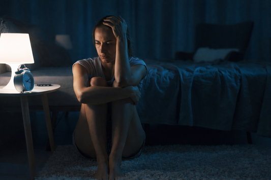 Woman sitting on the floor beside her bed at night, arms wrapped around her knees, looking down in quiet sadness while a lamp glows nearby.