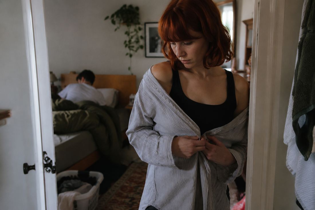 Red-haired woman covering herself with an oversized shirt while her partner sits on the bed, reflecting insecurity about being naked in a relationship.