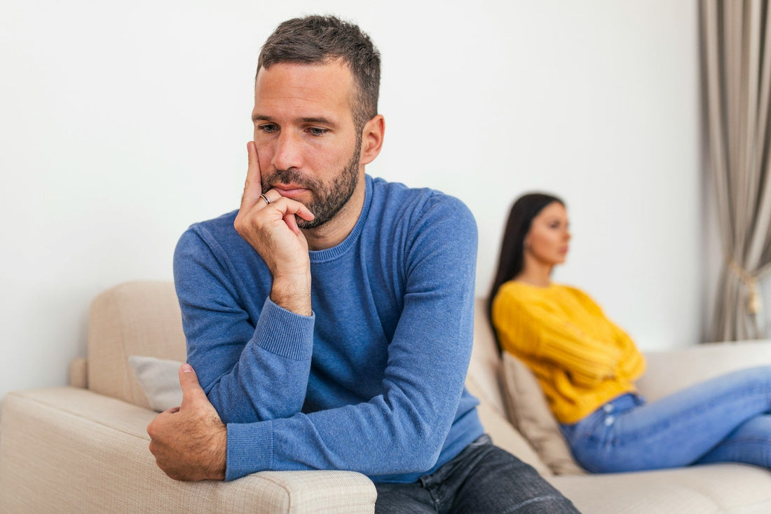 Man sitting apart from his partner after relationship conflict, looking distressed and emotionally withdrawn while she sits in the background.