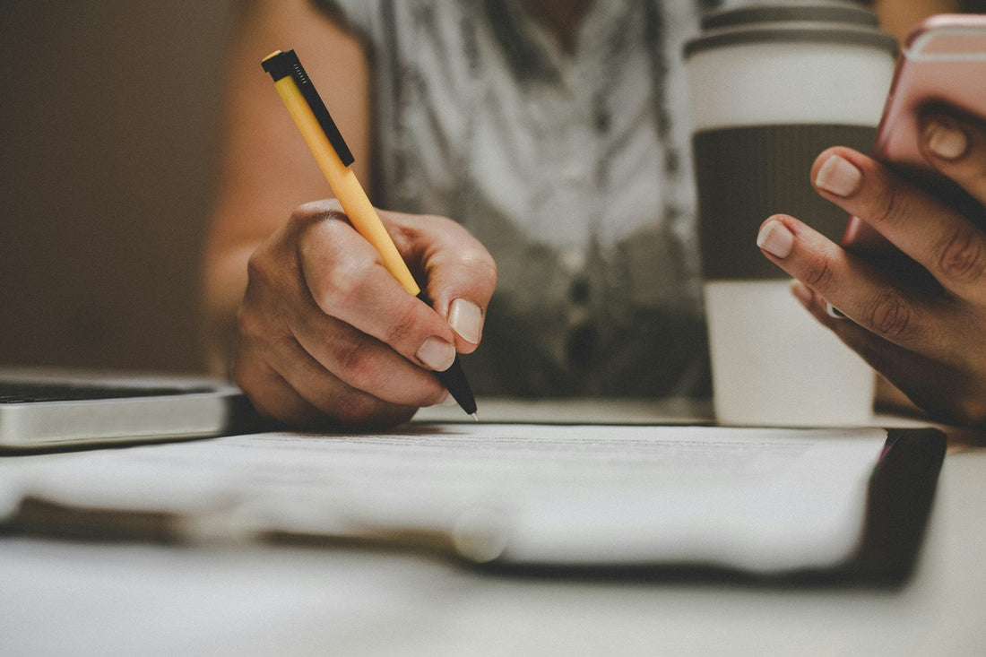 A close-up lifestyle shot of a person writing on a document with a yellow and black pen, holding a smartphone in the other hand, with a coffee cup in the background.