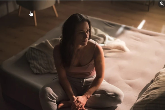 Woman sitting cross-legged on an unmade bed at night, looking off to the side while holding her hands together in quiet thought.