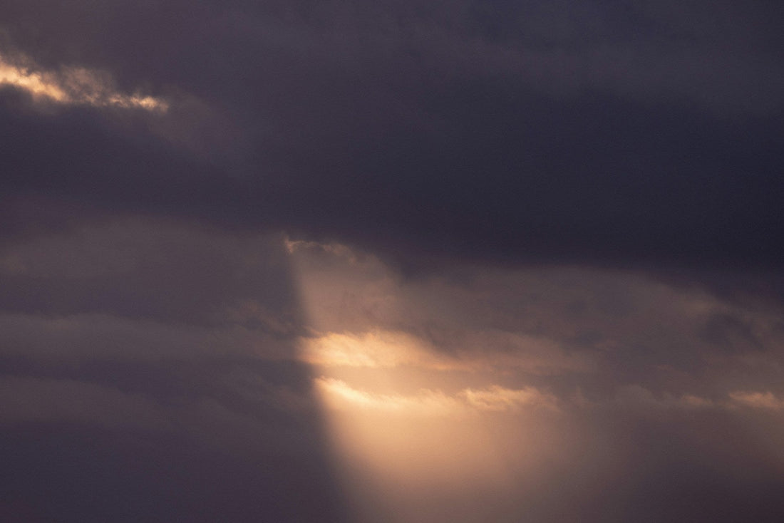 Dark storm clouds with a beam of sunlight breaking through, casting a soft glow of warm gold and orange light.