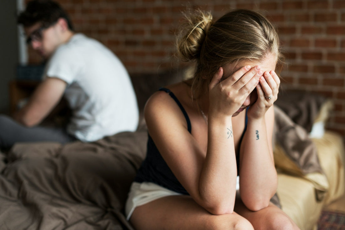 A woman sits on a bed in a bedroom, covering her face with her hands. An avoidant man is seated in the background on the same bed, turned away, with a brick wall behind them 