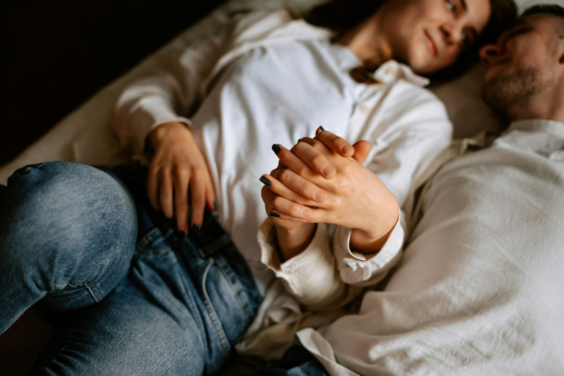 A couple lying close together on a bed, holding hands. The woman wears a white long-sleeve top and blue jeans, and the man wears a white shirt.