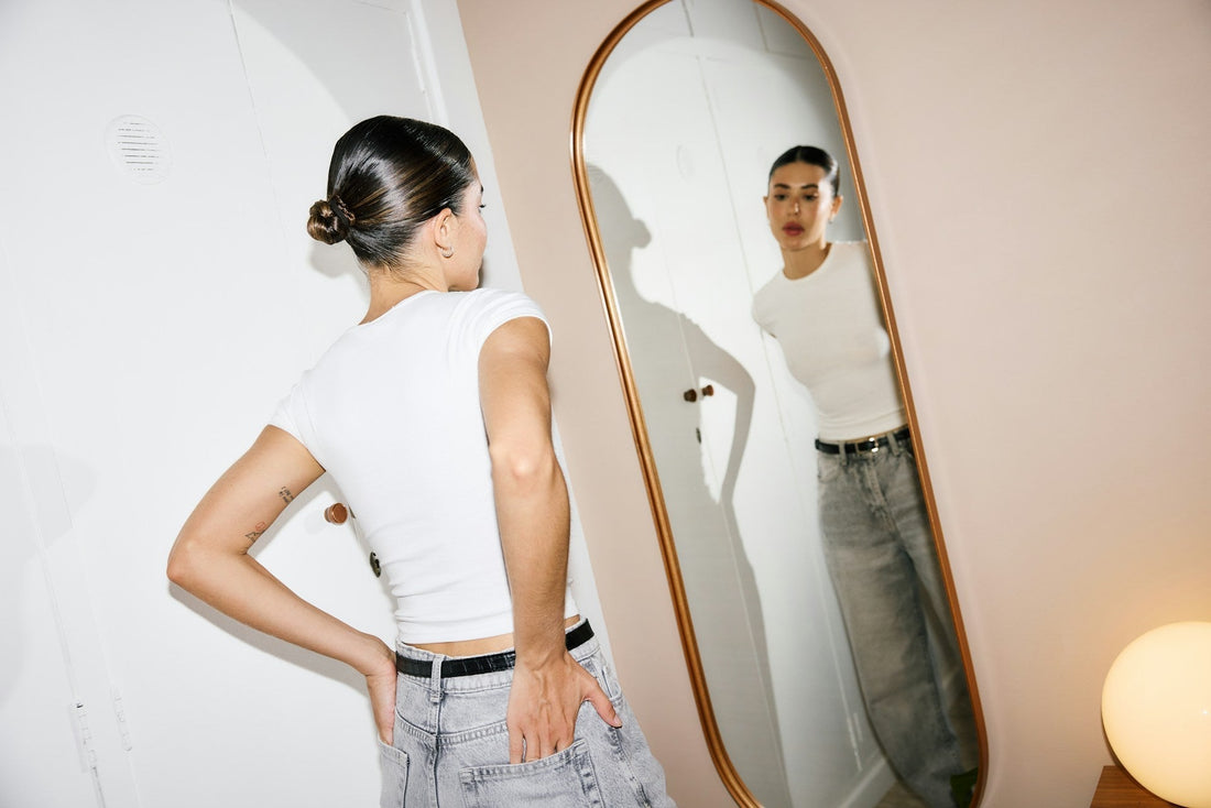 A woman wearing a white ribbed short-sleeve crop top and light-wash jeans stands in front of a large arched mirror, looking at her reflection, studying her reflection while comparing herself to someone new. 