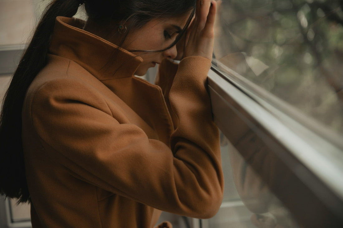 Woman standing by a window with her head lowered, reflecting feelings of self-doubt and minimizing what happened after a harmful relationship.