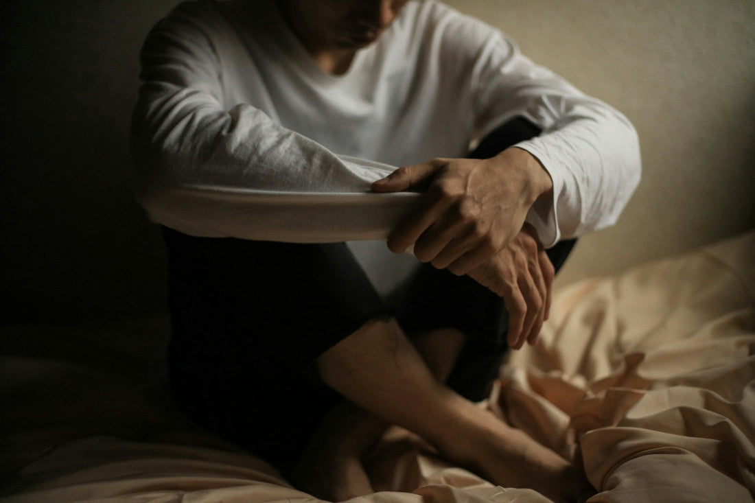 A person sitting curled up on a bed in a dimly lit room, wearing a white long-sleeve shirt and dark pants, with bare feet visible on rumpled bedding.