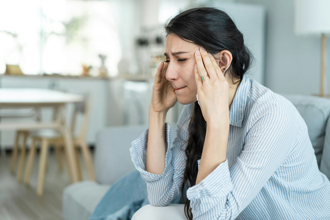 A woman with dark hair sits on a light gray couch in a bright living room, holding her temples in distress, replaying the last conversation with her ex