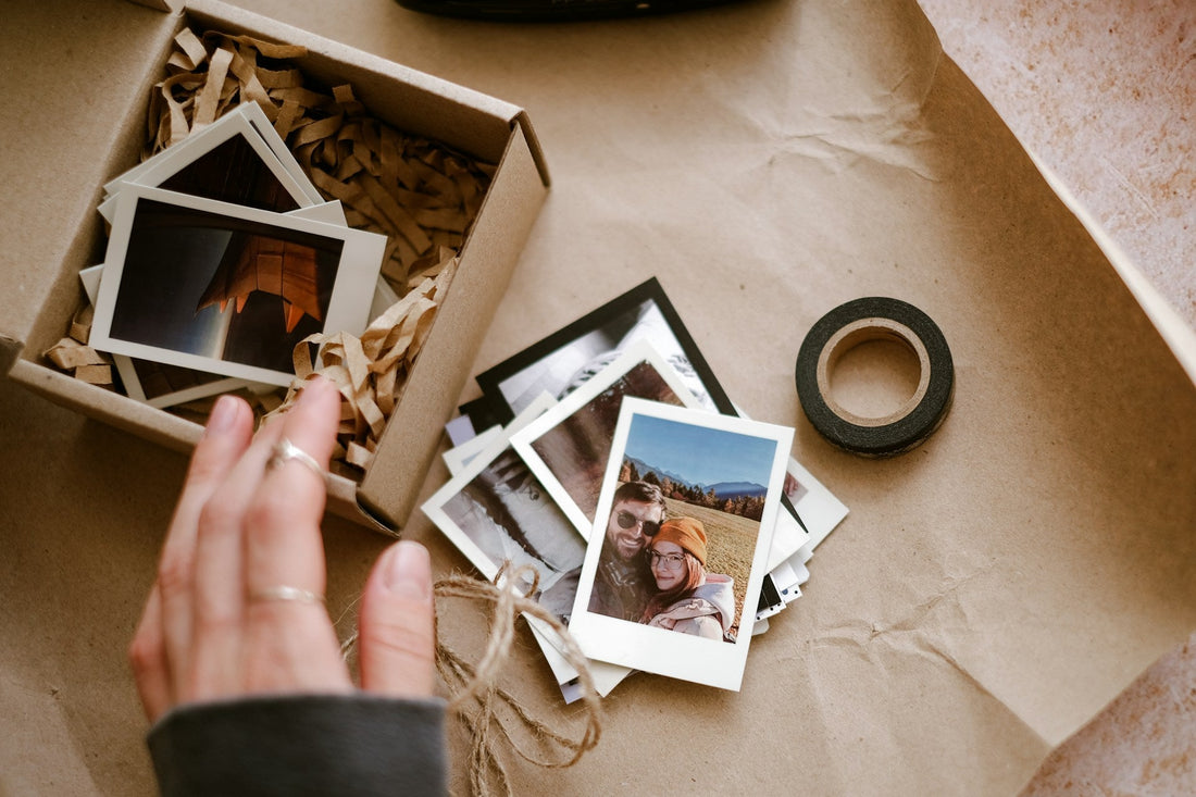 A hand is arranging instant photographs inside a kraft paper box with shredded paper filler, alongside a roll of black tape on a brown paper background.