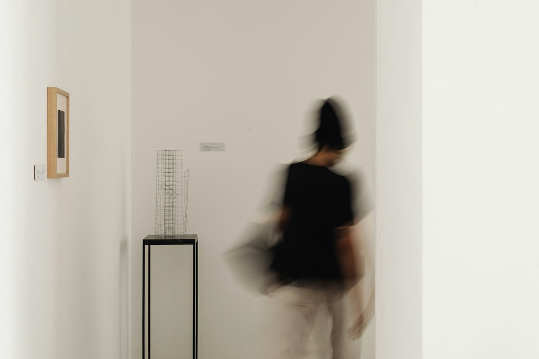 A blurred person in a black top and light pants walks through a minimalist white gallery hallway with framed wall art and a black display stand.