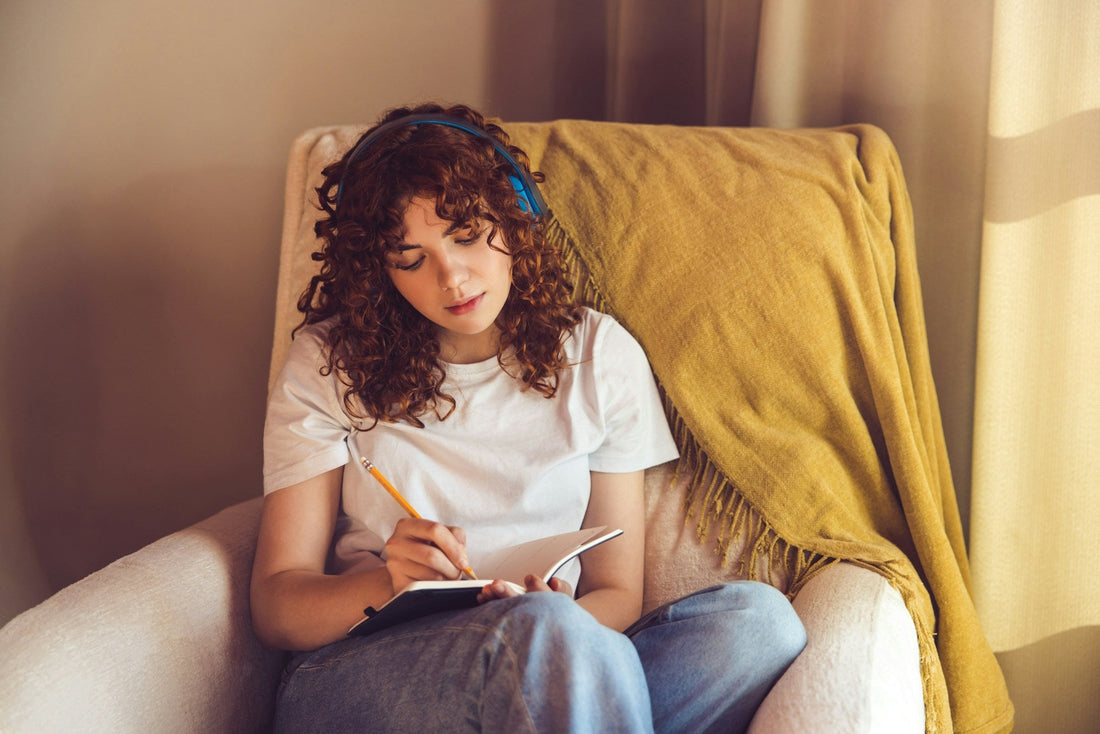 Woman wearing headphones and writing in a notebook while sitting in a chair, using journaling to process breakup feelings privately.