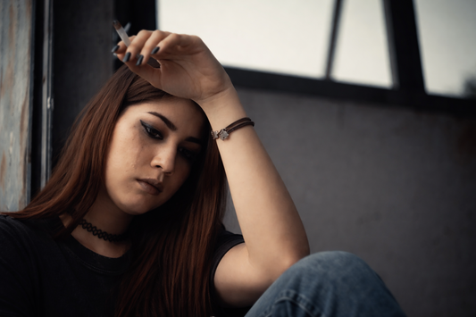 Young woman sitting by a window holding a cigarette, looking down with a distant expression, capturing the quiet aftermath of a breakup and thoughts left unsaid.