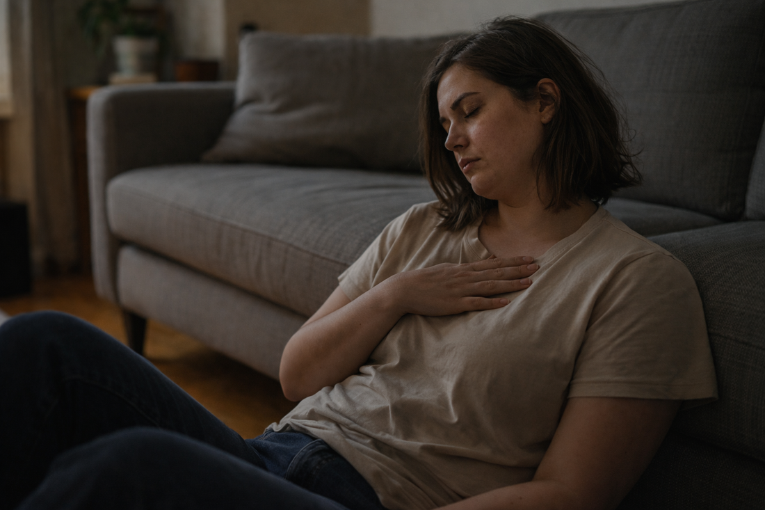 Woman sitting on the floor beside a couch in soft evening light, hand resting on her chest as she sits quietly with unspoken emotion.
