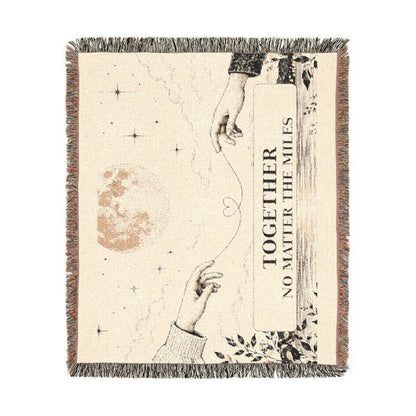 A square woven throw blanket with fringe edges, featuring a celestial design with stars, a moon, two reaching hands, and the text 'TOGETHER NO MATTER THE MILES' on a cream background.